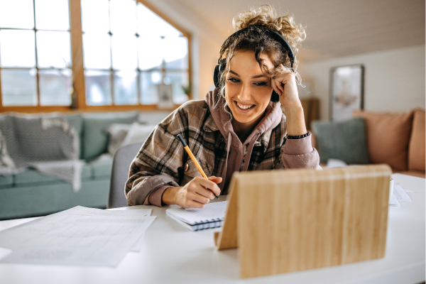 a teacher looking at her tablet screen, smiling, wearing headphones, with a pencil in hand ready to write in her notebook.